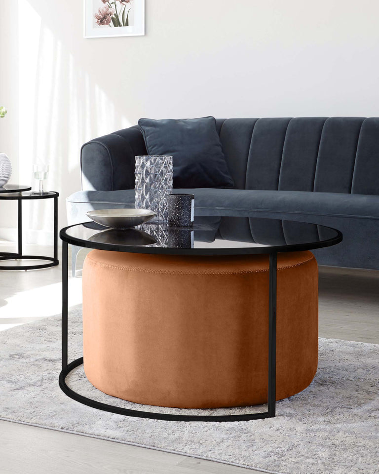 Elegant modern living room setup featuring a sleek dark grey velvet sofa with vertical tufting and two round nested coffee tables with black metal frames; the larger table has a black glass top, and the smaller table has a warm caramel-coloured leather upholstery. The set is placed on a textured light grey rug over a light wooden floor, complementing the minimalist décor.