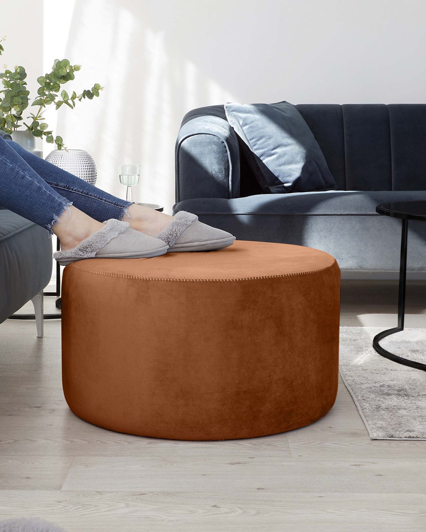 A tan round ottoman in a velvety fabric is positioned in front of a charcoal grey fabric sofa adorned with blue-grey decorative pillows. A round black side table and a light grey area rug accent the modern living space.