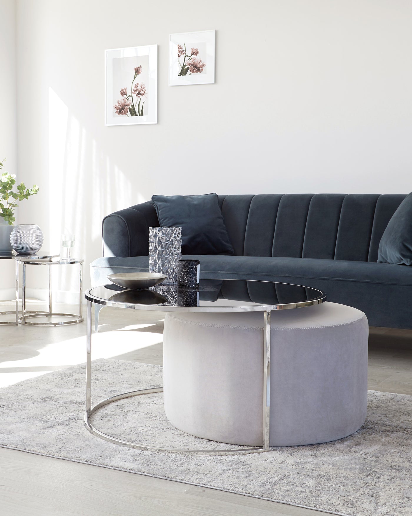 Elegant living room setup featuring a modern navy blue velvet sofa with tufted backrest, a round glass-top coffee table with a unique silver metal base, and a complementary light grey upholstered round ottoman. The room is accented with a textured light grey area rug, two framed art pieces on the wall, and a small silver side table with decorative items.
