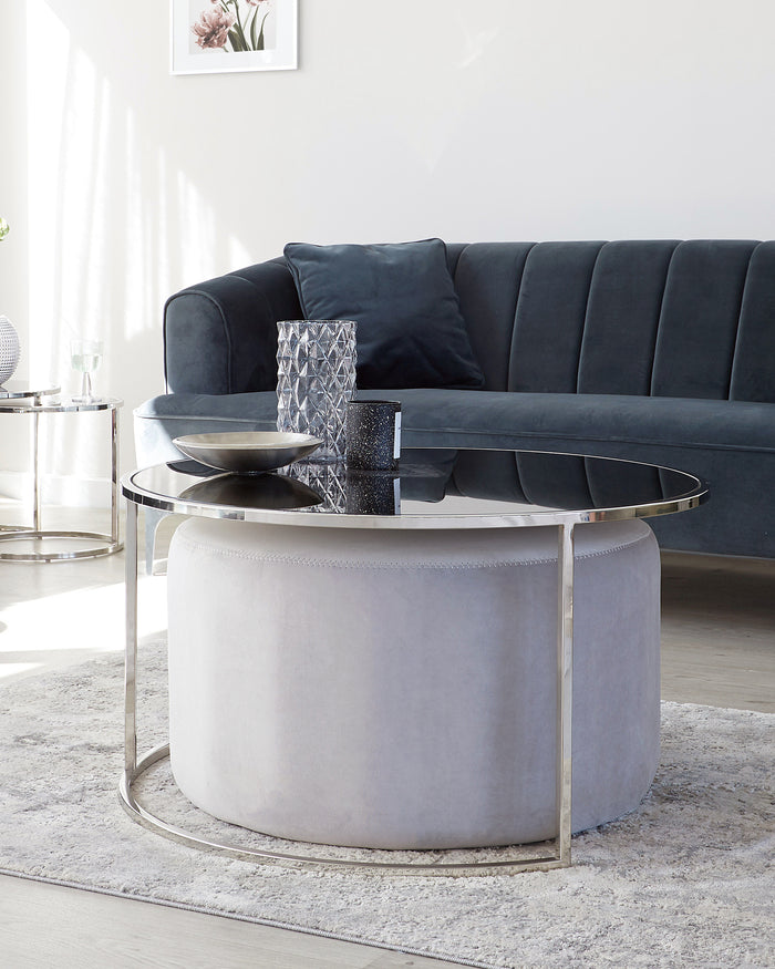 Elegant living room setup featuring a modern, deep blue velvet sofa with clean lines and plush seating, accompanied by a round, chic ottoman with a soft, light grey fabric cover and a unique, sleek circular metal base. In front of the sofa, there's a contemporary round coffee table with a reflective glass top and a silver metallic frame, enhancing the luxurious aesthetic of the space.