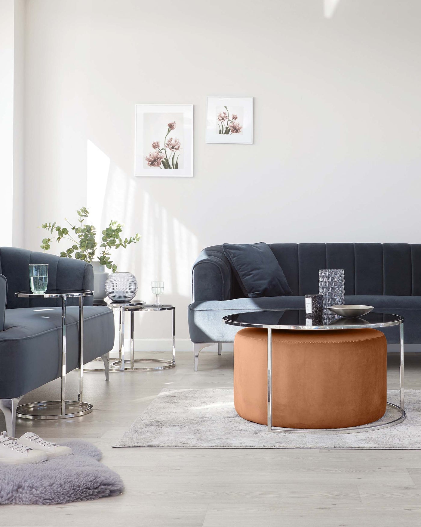 Modern living room arrangement featuring a dark blue velvet sectional sofa with tufted backrest, a circular caramel-coloured ottoman with a glass table top centrepiece, two round side tables with glass tops and silver metallic bases, a plush grey area rug beneath the furniture, and decorative accent pieces including pillows, a vase, and a silver textured sphere.