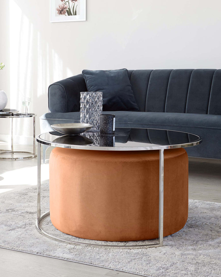 Contemporary living room furniture, featuring a dark grey velvet sofa with plush cushions and a round, tan leather ottoman with metallic silver base. In front of the sofa is a modern round coffee table with a glass top and silver frame.