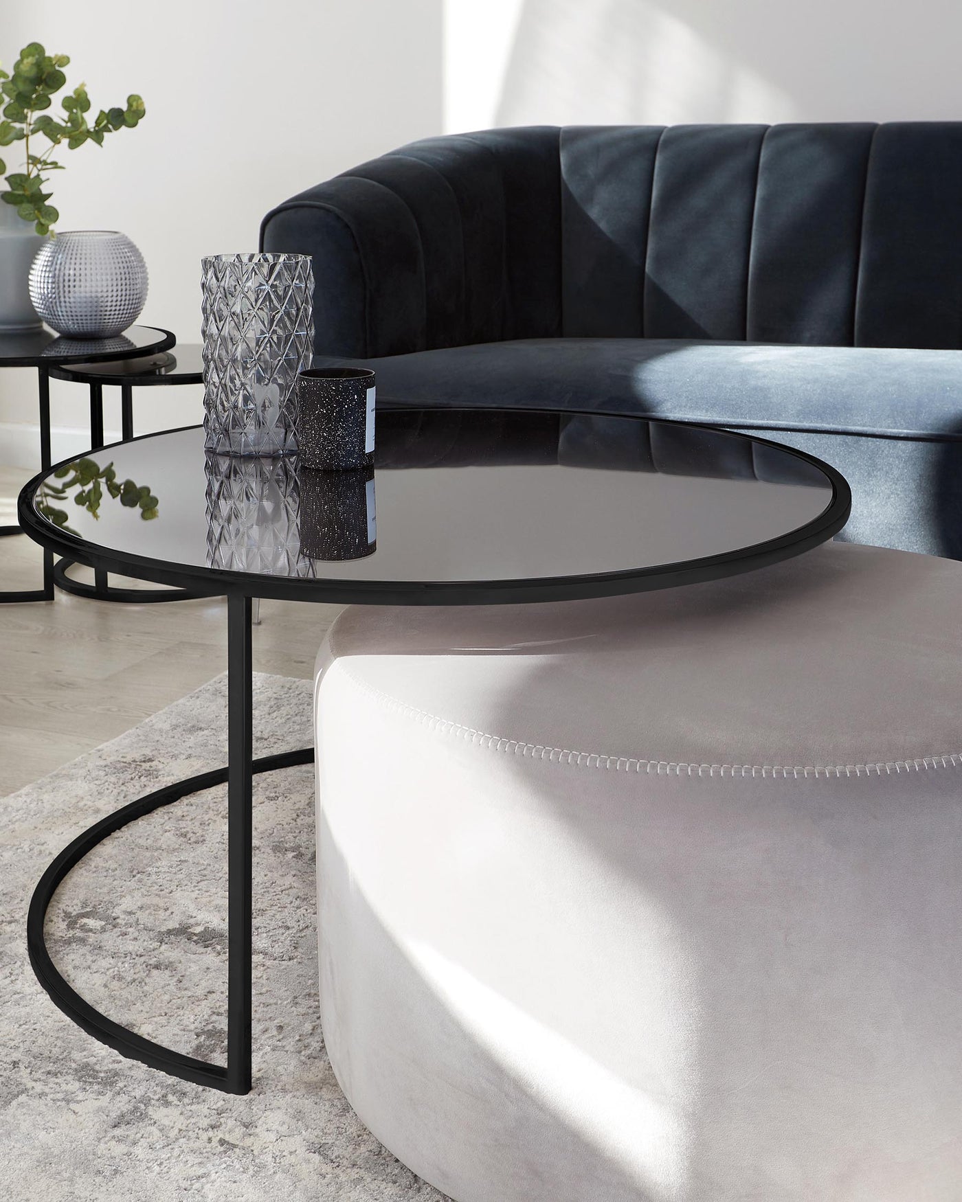 Elegant contemporary living room furniture featuring a sleek black velvet curved sofa, a round glass-top side table with a black metal frame, and a light beige upholstered round ottoman with detailed stitching.