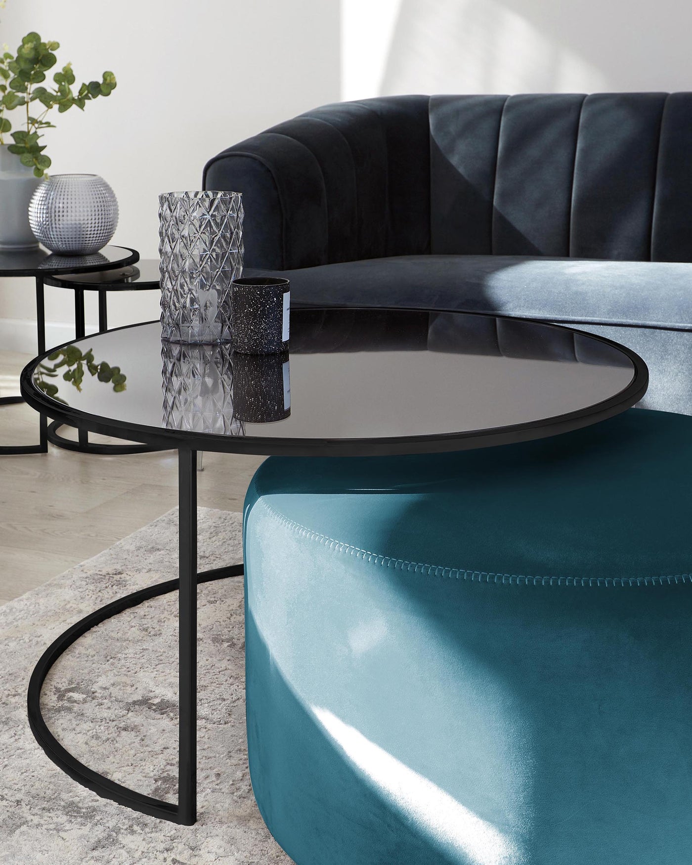 Elegant living room scene featuring a plush, curved dark grey velvet sofa, a round, teal ottoman, and a pair of circular black metal side tables with glossy black tops, accented with decorative vases.