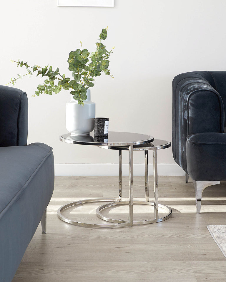 Modern furniture setting featuring a sleek round side table with a chromed metal base and a clear glass top, positioned between two plush velvet armchairs in a deep blue colour. The armchairs have a contemporary design with rounded backs and comfortably cushioned seats, providing a sophisticated look suitable for a chic living space. The table is accessorised with a white vase holding greenery and a black decorative candle, enhancing the elegant ambiance of the arrangement.