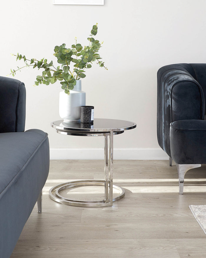 Modern round side table with a chrome-finished metal frame and a sleek glass top, accompanied by a dark upholstered armchair with velvet fabric and silver leg accents.