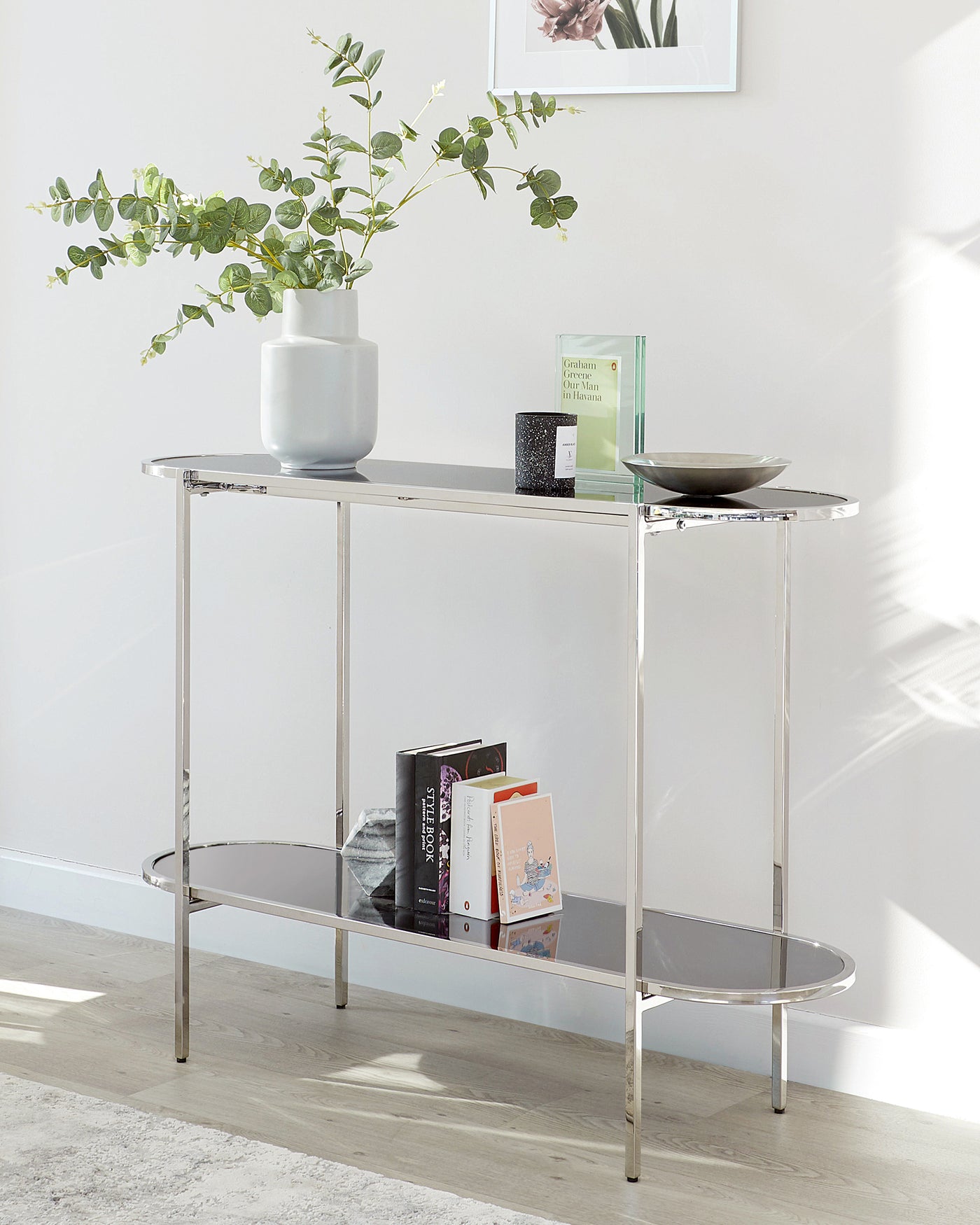 A modern, sleek console table with a reflective chrome finish and two levels. The top surface is rectangular, while the lower shelf is semi-circular, creating an elegant contrast. The table is adorned with a vase of greenery, books, and decorative items.