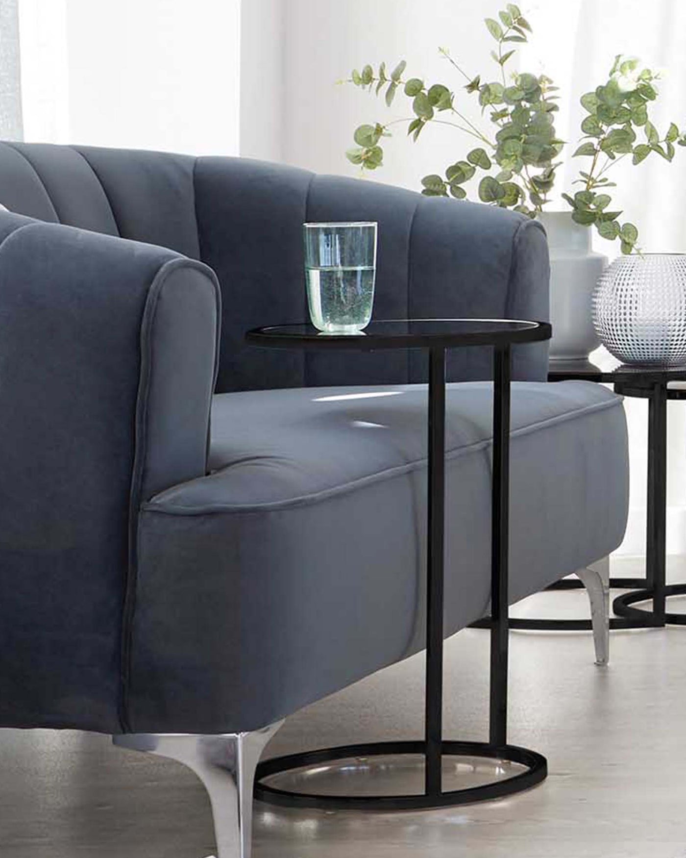 Elegant modern dark grey sofa with vertical channel tufting and sleek metallic silver legs, alongside a round black side table with a glass top and metal base, featuring a cylindrical clear glass vase with water.