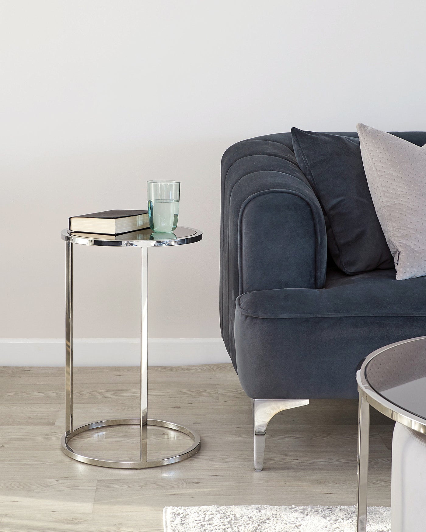 A modern, round, glass-top side table with a chrome base, positioned next to a dark grey fabric upholstered sofa with plush cushions.