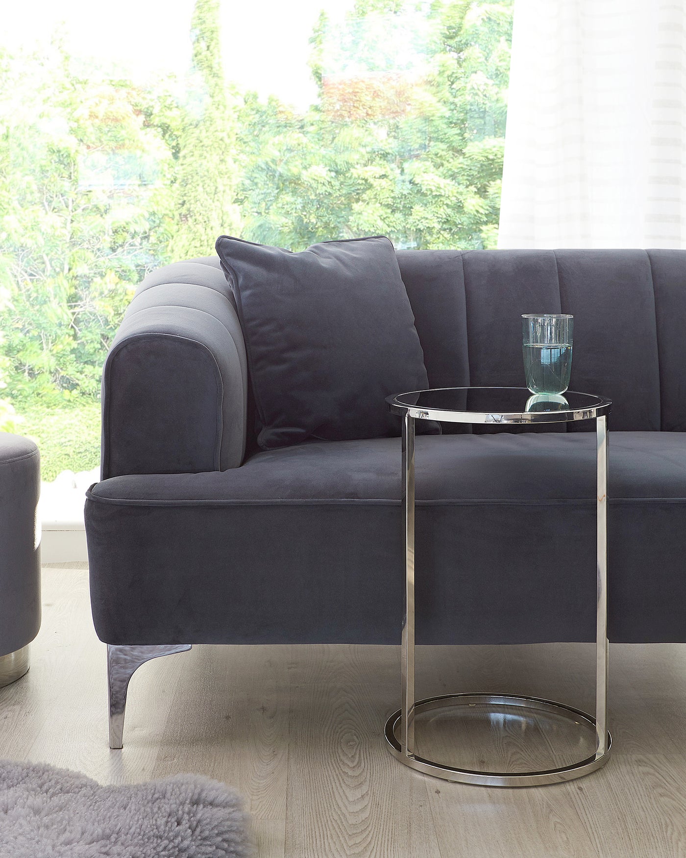 Contemporary dark grey fabric sofa with plush cushions and a round, chrome accent side table with a clear glass top.