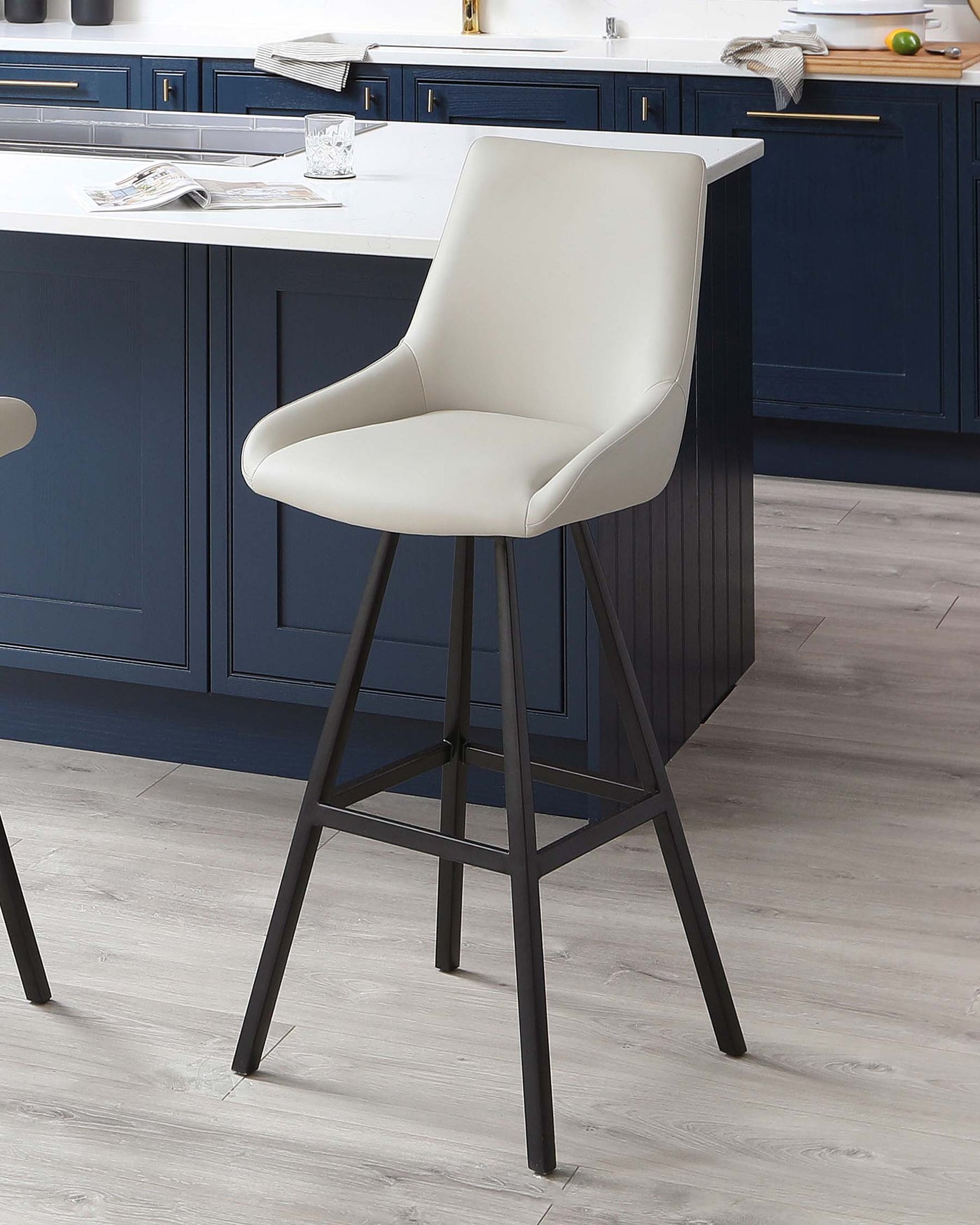A modern bar stool with a high back and armrests, upholstered in light beige faux leather, set on a tall, angular, black metal base with four legs and supporting footrests. The stool is set against a kitchen island with navy blue cabinetry and a white countertop.