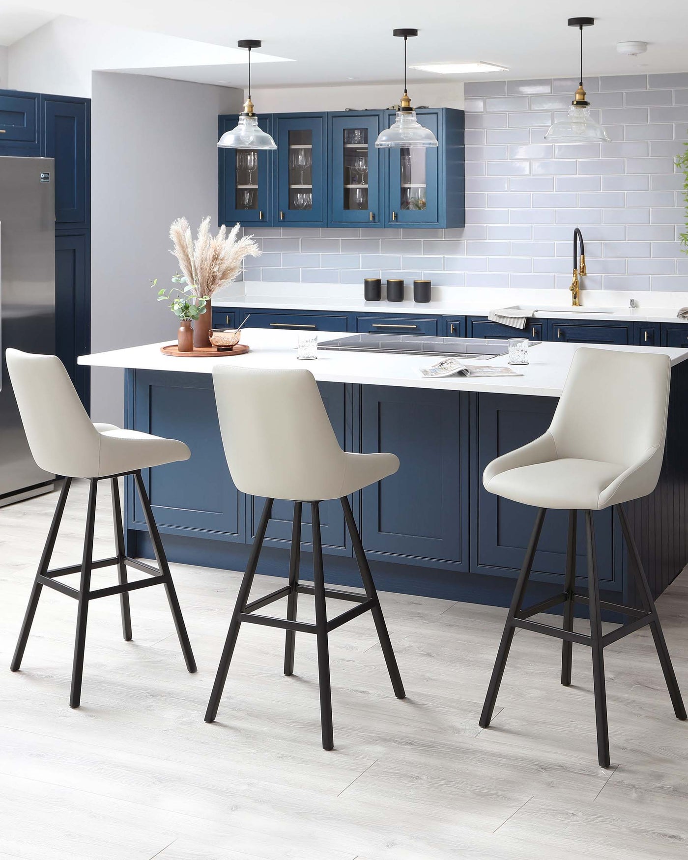 Three modern bar stools with beige upholstered seats and black, angled metal legs positioned at a white kitchen island.