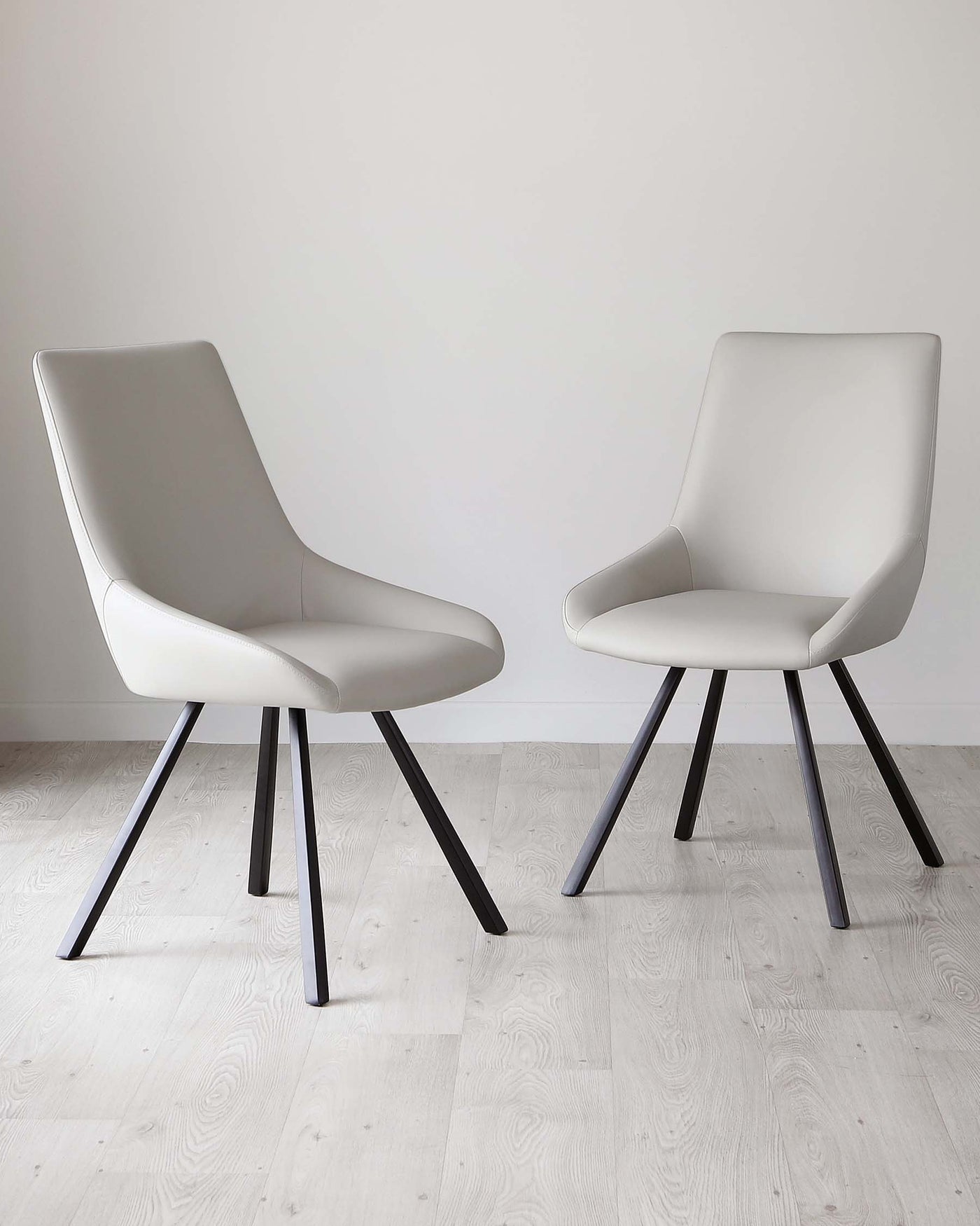 Two modern dining chairs with sleek, light grey upholstery and black metal legs, positioned on a herringbone-patterned light wooden floor against a clean white background.