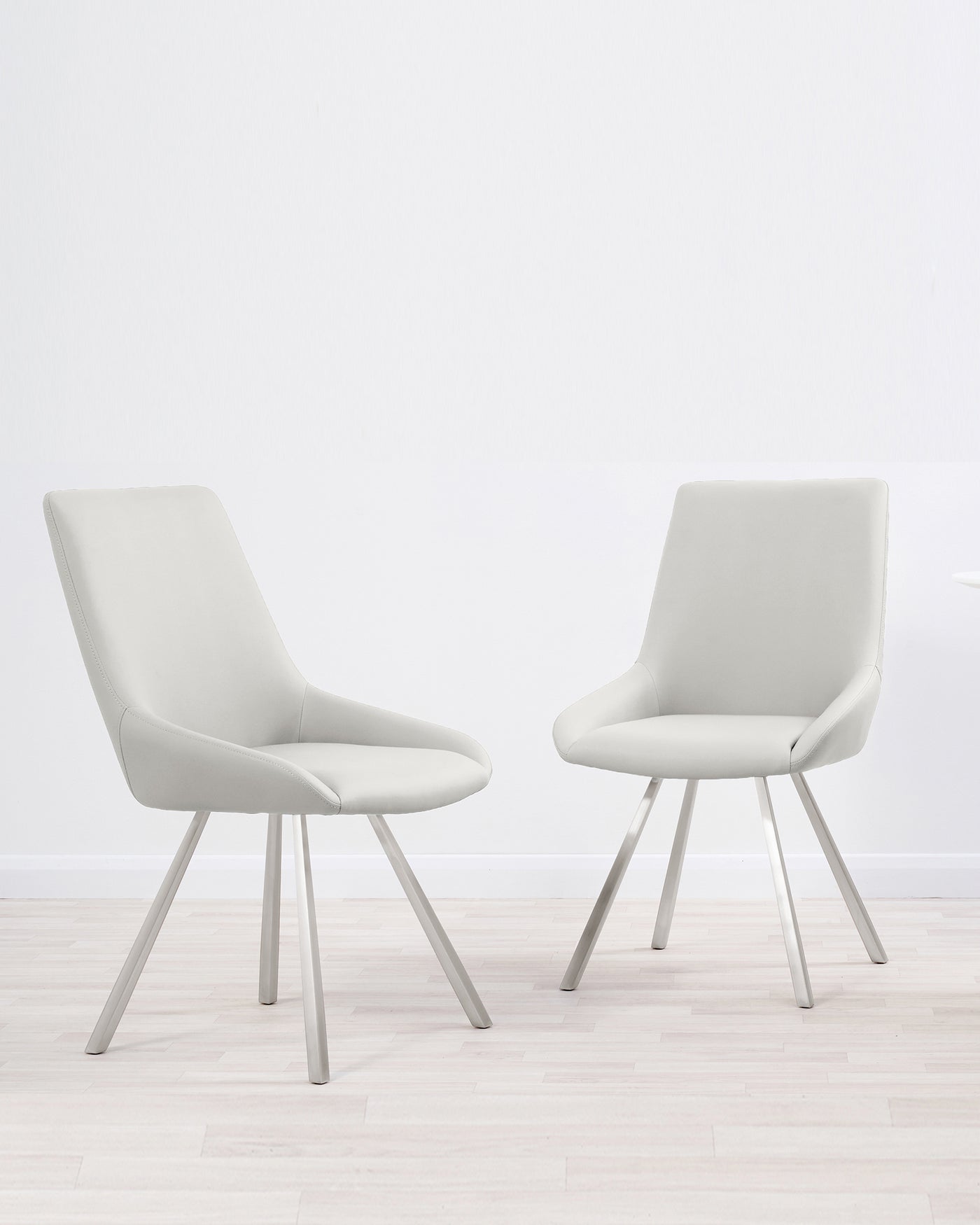 Two modern light grey upholstered dining chairs with a sleek design and splayed metal legs, set on a light hardwood floor against a white wall.