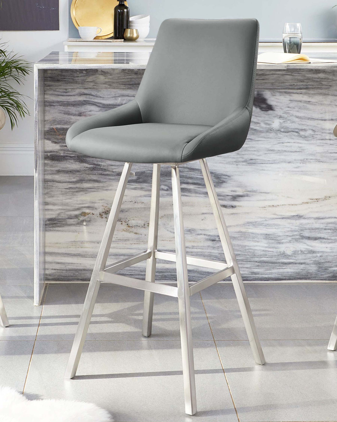 Modern grey upholstered bar stool with a curved backrest and sleek silver metal legs, set against a marble counter backdrop.