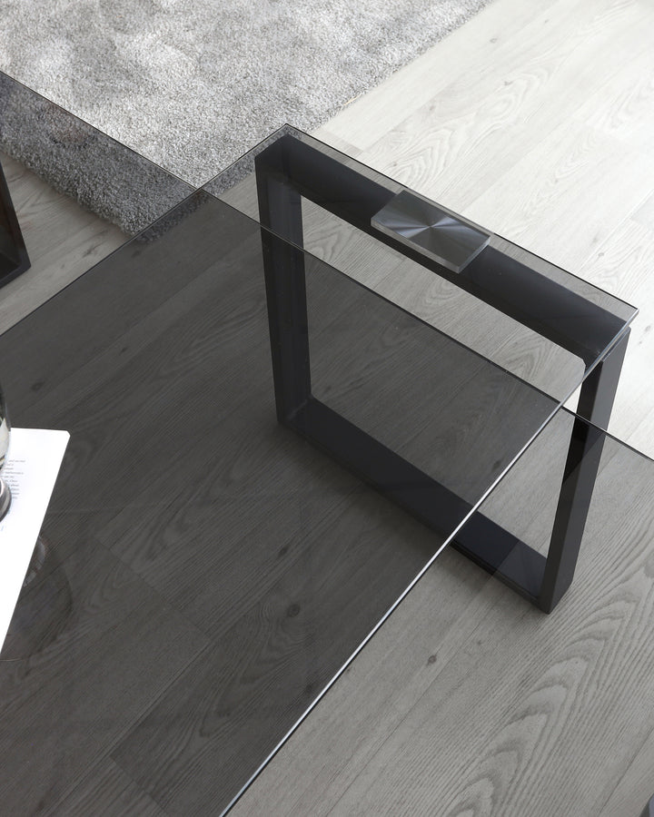 Modern glass-top coffee table with a unique black geometric metal base on a hardwood floor, adjacent to a plush grey area rug.