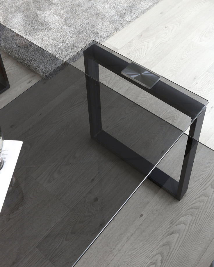 Modern glass-top coffee table with a unique black geometric metal base on a hardwood floor, adjacent to a plush grey area rug.