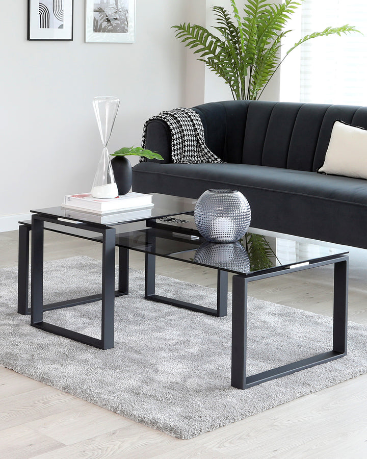 A set of two contemporary nested coffee tables with black metal frames and clear glass tops, positioned on a light grey shaggy rug in a living room setting. The larger table has decorative items on it, including a large white hourglass, books, and a small vase. A modern black sofa with vertical channel tufting and a houndstooth throw blanket is partially visible in the background.