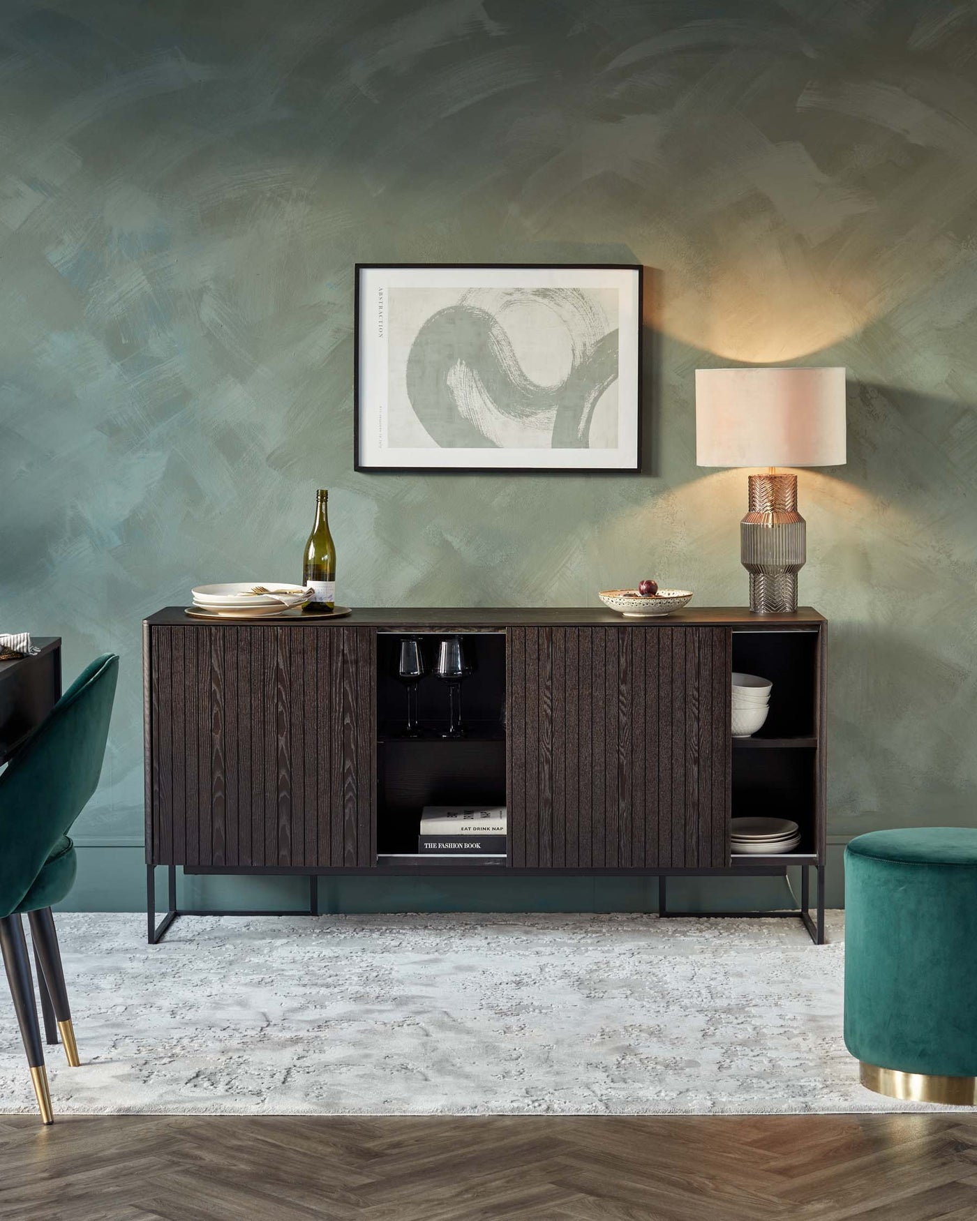 Elegant modern furniture in a stylish interior, featuring a dark wooden sideboard with a textured front, metal legs, and open shelves displaying dishware and books. A velvet green ottoman adds a pop of colour, while a sophisticated table lamp with a cylindrical base and a white shade provides warm lighting. The decor is completed with a framed abstract art piece above the sideboard, creating a cohesive and refined aesthetic.