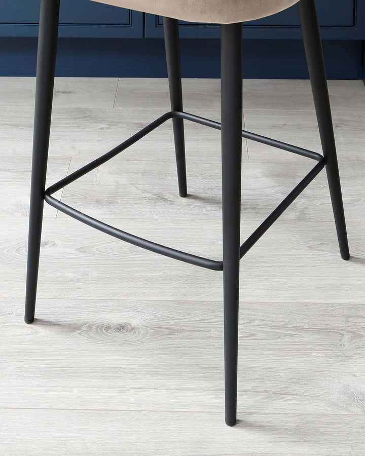 Modern minimalist stool with a round, light beige seat and sleek, black metal legs featuring geometric stability bars. The stool is set against a light wood laminate floor with a blue-grey wall in the background.
