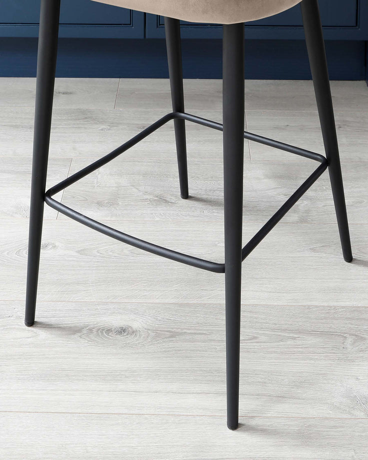 Modern minimalist stool with a round, light beige seat and sleek, black metal legs featuring geometric stability bars. The stool is set against a light wood laminate floor with a blue-grey wall in the background.