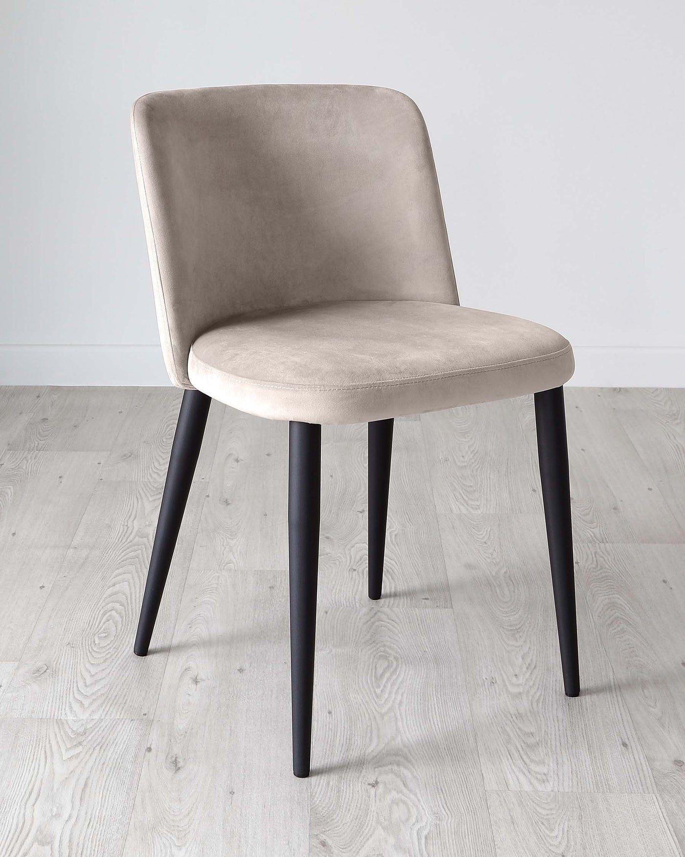 Modern chair with a taupe velvet upholstered curved backrest and seat, supported by four sleek black metal legs, positioned on a light wooden floor against a white wall background.