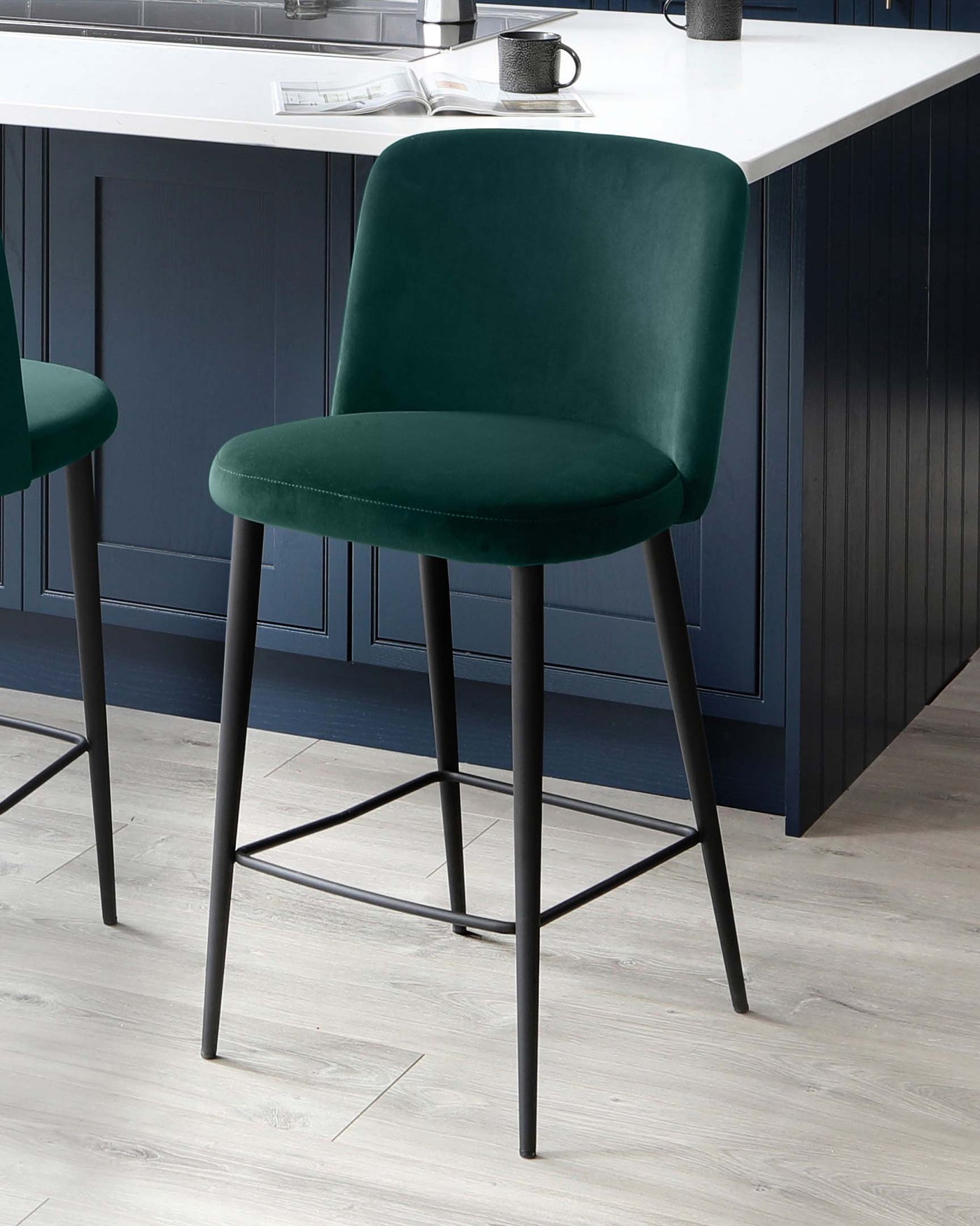 Elegant modern bar stools with luxurious dark green velvet upholstery and sleek black metal legs, featuring a minimalist backrest and footrest, showcased in a stylish kitchen with a white countertop and dark blue cabinetry.