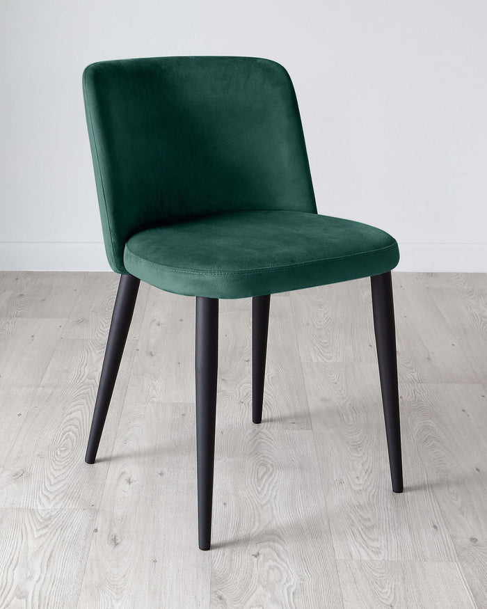 Elegant modern chair with plush dark green velvet upholstery and angular black metal legs, featuring a curved backrest and a comfortable padded seat, set against a light hardwood floor and a white wall backdrop.
