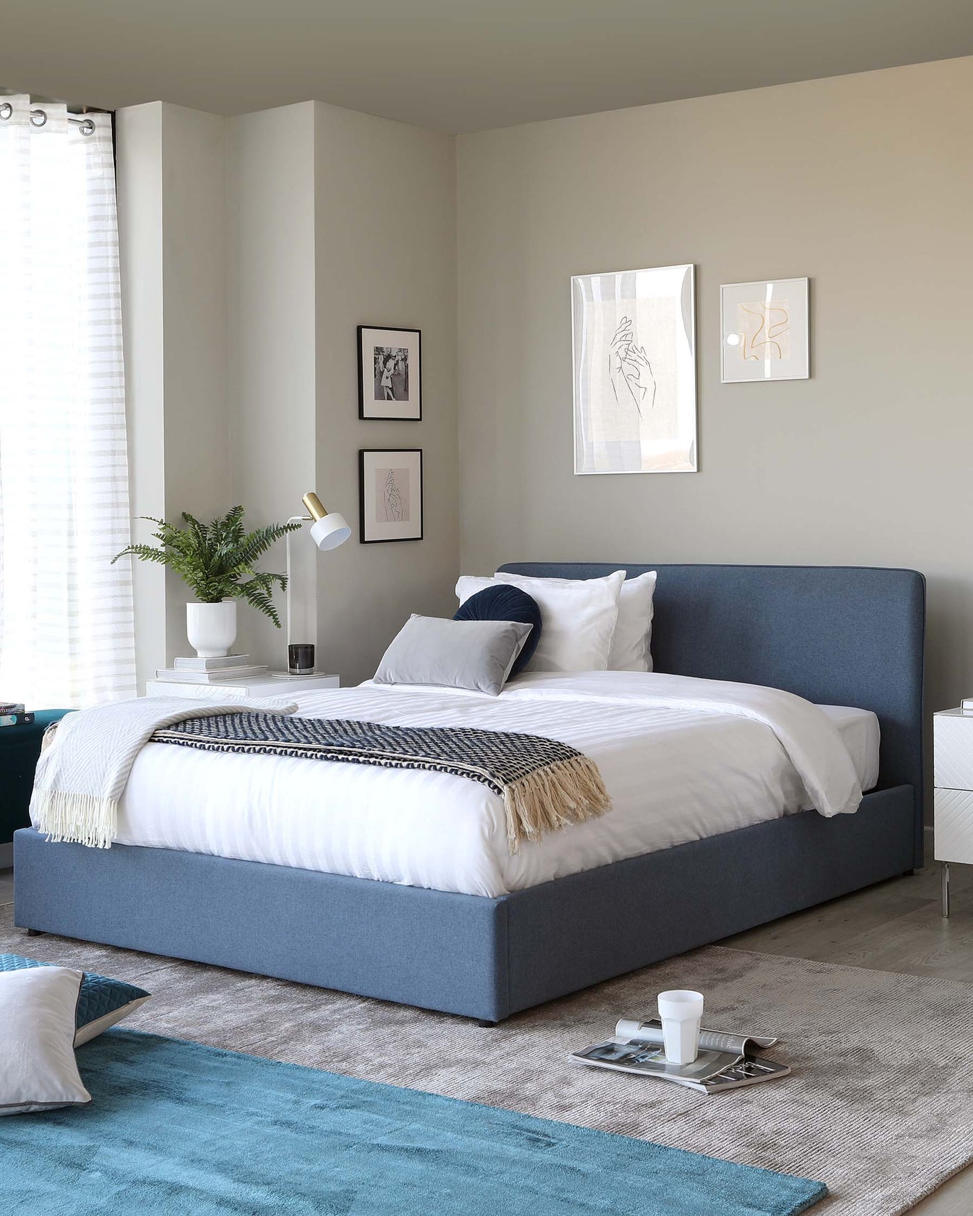 A modern bedroom setting featuring a queen-sized bed with a minimalist blue upholstered headboard and platform base. The bed is adorned with crisp white bedding, a patterned throw, and assorted pillows. A white side table with a simple design and round white lamp provides a functional accent to the space. A plush area rug in a blend of neutral and teal hues adds warmth and texture to the floor.