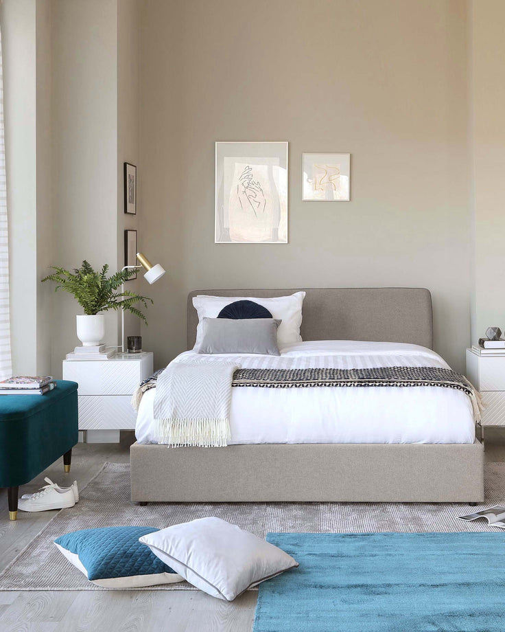 A modern bedroom setup with a grey upholstered bed featuring a tufted headboard, flanked by two white nightstands with clean lines. One nightstand has a white vase with greenery, the other is partially visible with a closed drawer. A bench upholstered in a rich teal fabric sits at the end of the bed, adding a pop of colour. Completing the scene, a plush light grey area rug is overlaid with a smaller blue accent rug, which provides additional texture and colour contrast to the room.