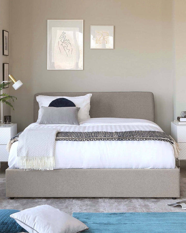 Modern minimalist bedroom featuring a queen-sized bed with a tailored upholstered headboard in a soft grey fabric, flanked by two white bedside tables with matching white contemporary lamps. A textured throw blanket in black and white complements white bed linens, while a large pillow in navy blue adds a pop of colour. The scene is anchored on a light grey area rug with subtle blue accents, contributing to the calm and serene palette of the room.