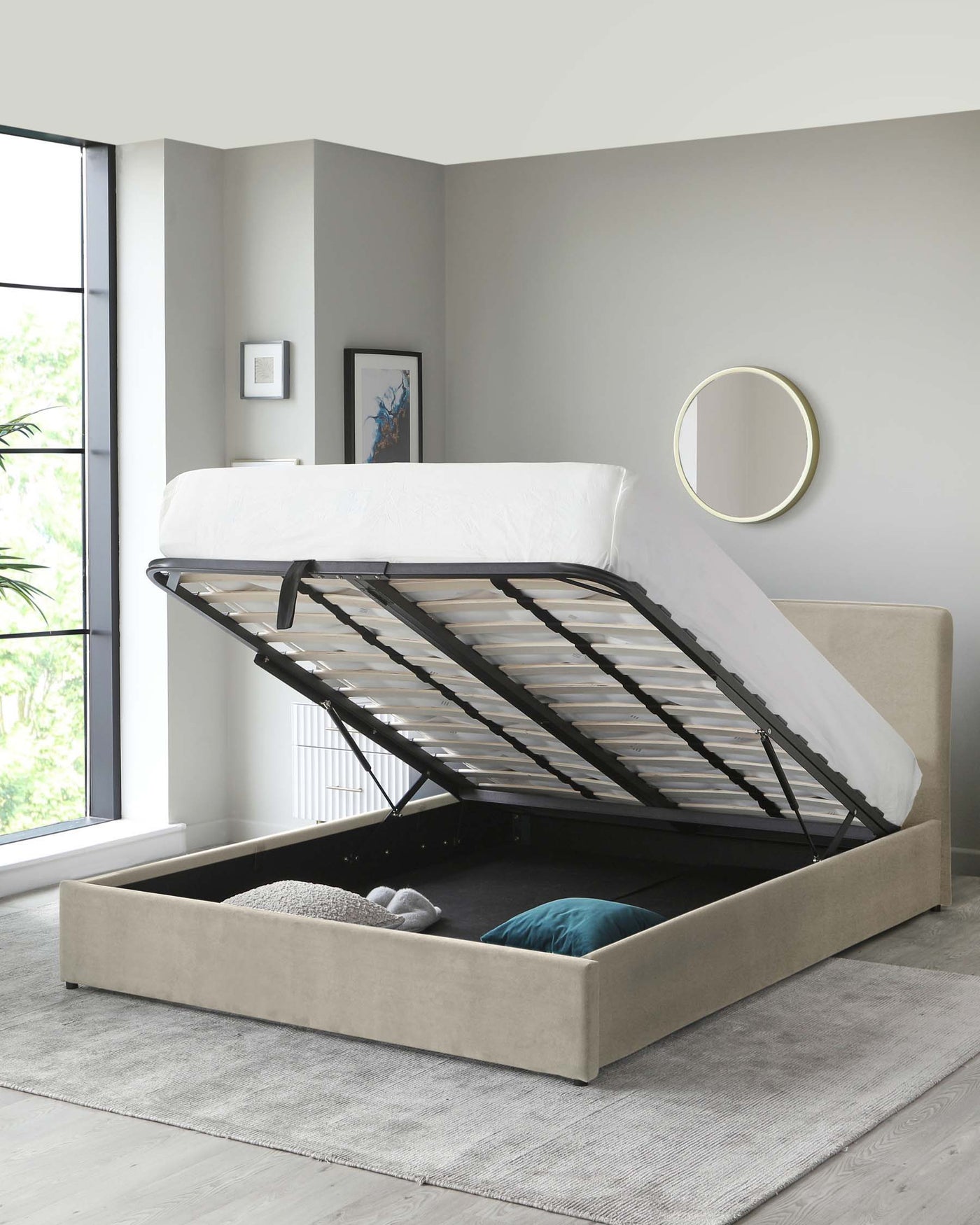 Modern beige upholstered storage bed with a lifted mattress frame revealing under-bed storage space containing pillows and blankets, situated on a light grey area rug. A round mirror is mounted on the wall above the bed.