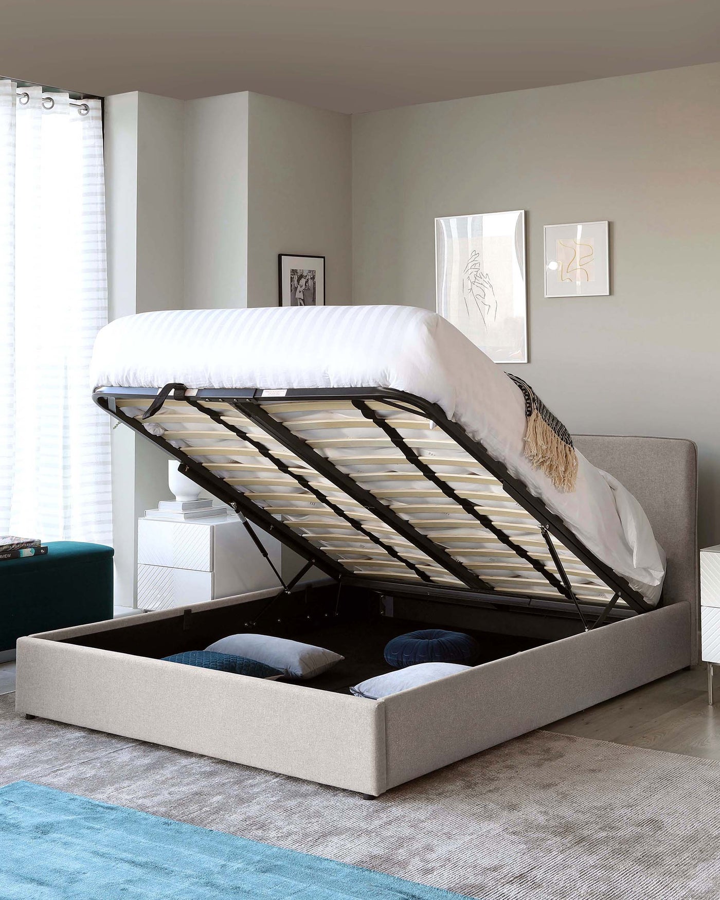 Modern upholstered storage bed with a lifting frame, showcasing a spacious under-bed storage compartment. The bed features a light grey fabric finish, a cushioned headboard, and a slatted base for mattress support.