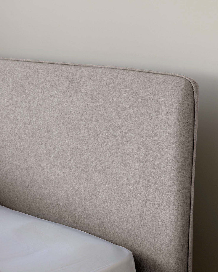 A close-up view of a contemporary upholstered headboard with a neutral heathered fabric finish, featuring a slightly curved top edge and clean, simple lines, suggesting a minimalist and modern aesthetic. The bedding or mattress is partially visible with a clean, plain white cover.