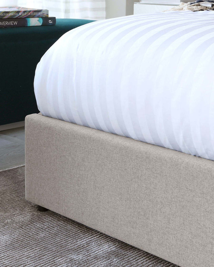 A contemporary beige upholstered bed frame with a low-profile footboard, complemented by an emerald green velvet tufted headboard in the background. A white striped duvet cover is partially visible, highlighting the bed's plush comfort. There's a hint of books suggesting a reading nook, thus presenting the furniture as part of a cosy, stylish bedroom setting.