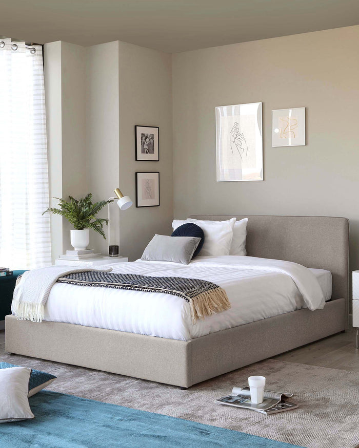 Modern minimalist style bedroom featuring a king-size platform bed with a padded, upholstered headboard in a neutral grey fabric. The bed is adorned with white and navy blue pillows and a complementary throw blanket with fringe detailing. A white nightstand with clean lines holds a simple white vase with green foliage, flanking one side of the bed. The décor is completed with a low-profile, grey and blue area rug under the bed, and a small, sleek white table lamp with a gold accent on the headboard.