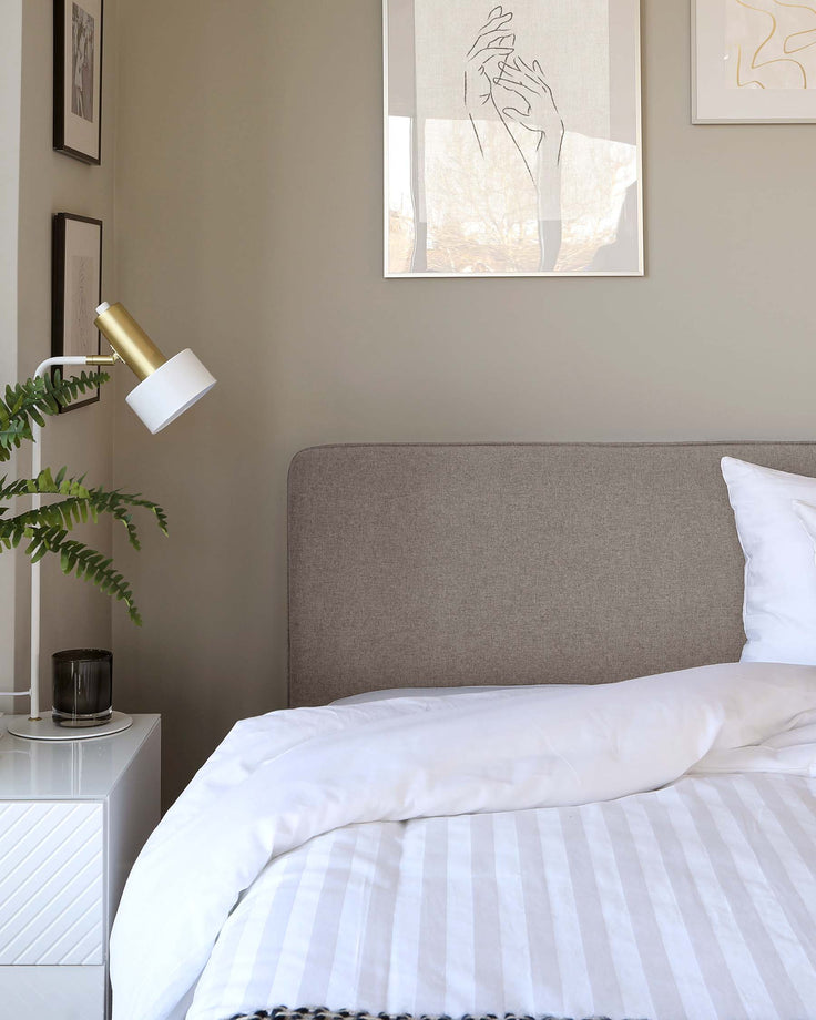 Modern bedroom featuring a neutral-toned upholstered headboard and a white nightstand with a reflective facade. The bedding is white with subtle stripes, and a decorative black-bordered throw blanket is placed at the foot of the bed. A sleek, white and gold wall-mounted lamp provides illumination.
