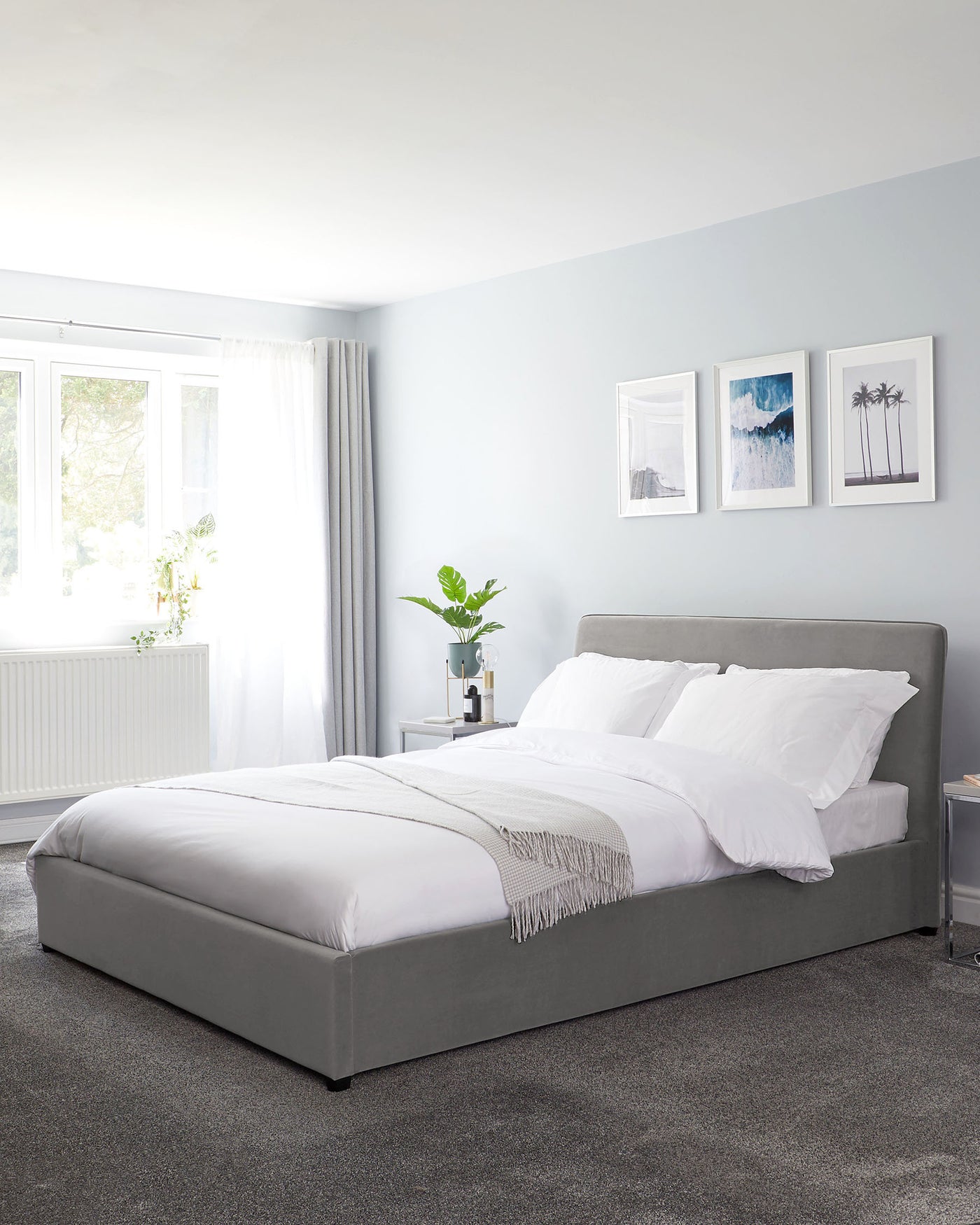 Elegant contemporary bedroom featuring a grey upholstered platform bed with a high headboard, dressed in white bedding and accented with a light fringe throw. The room has a simple bedside table with a potted plant to the side, set against a serene pale blue wall and plush grey carpeting.