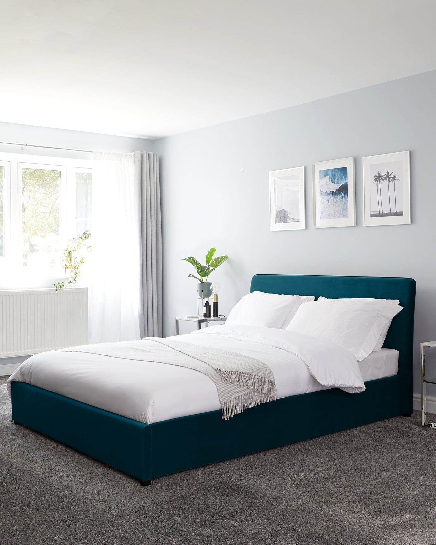 A modern bedroom featuring a plush teal upholstered bed frame with a high, curved headboard and a low-profile footboard. The bed is dressed in crisp white bedding with a contrasting light-grey fringed throw at the foot. The clean lines and bold colour of the bed make it a statement piece in the minimalist-styled room.
