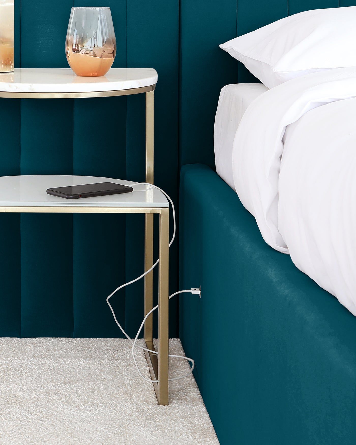Elegant modern bedside table with a marble-like top and gold-coloured metal frame, next to a plush teal upholstered bed with white bedding. A decorative glass vase rests on the table's upper tier.