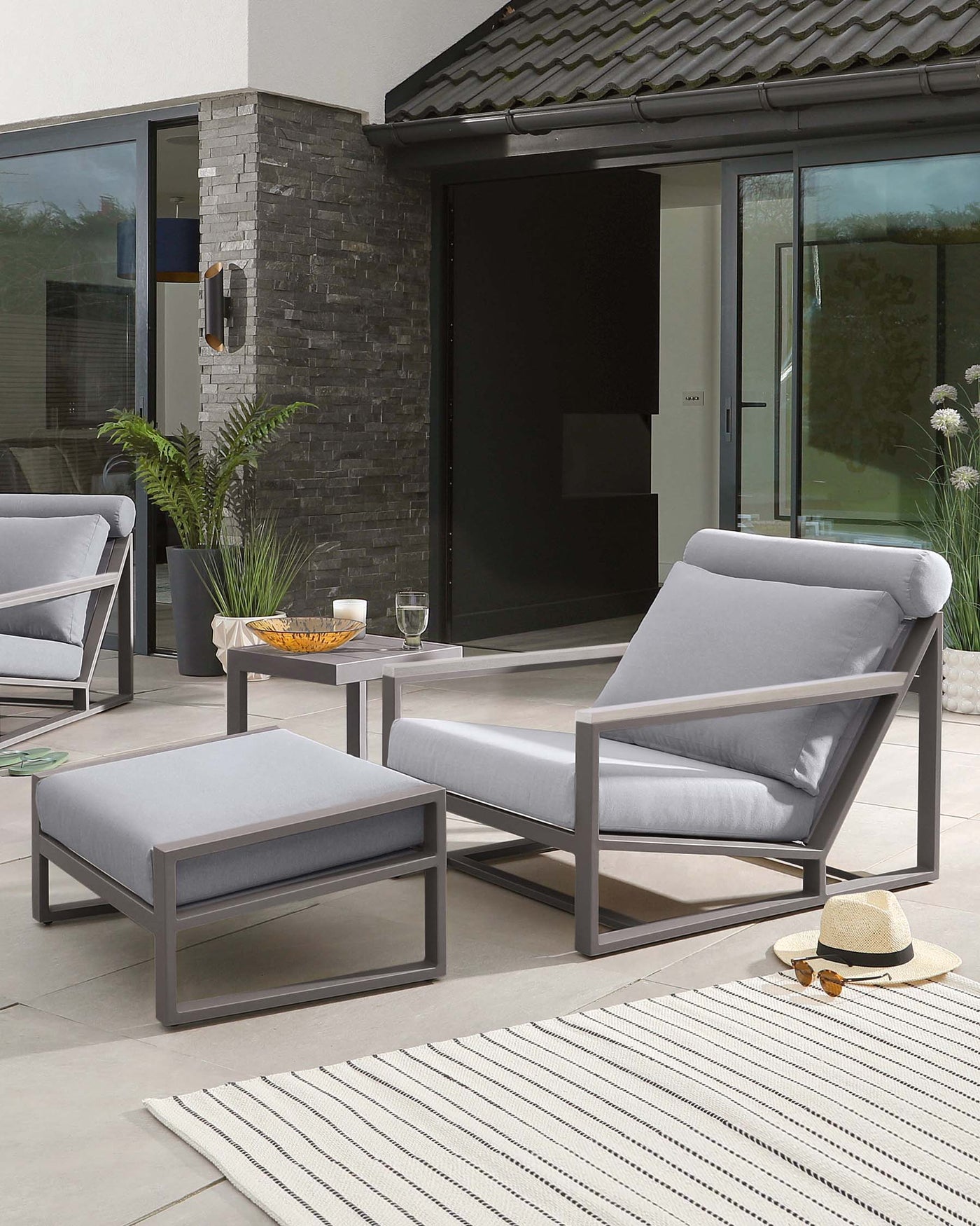 Modern outdoor patio furniture featuring a sleek grey metal frame with complementary light grey cushions. The set includes a comfortable armchair, a matching ottoman, and a simple, elegant side table with a clear tabletop. The design is clean and contemporary, perfect for chic outdoor lounging areas.