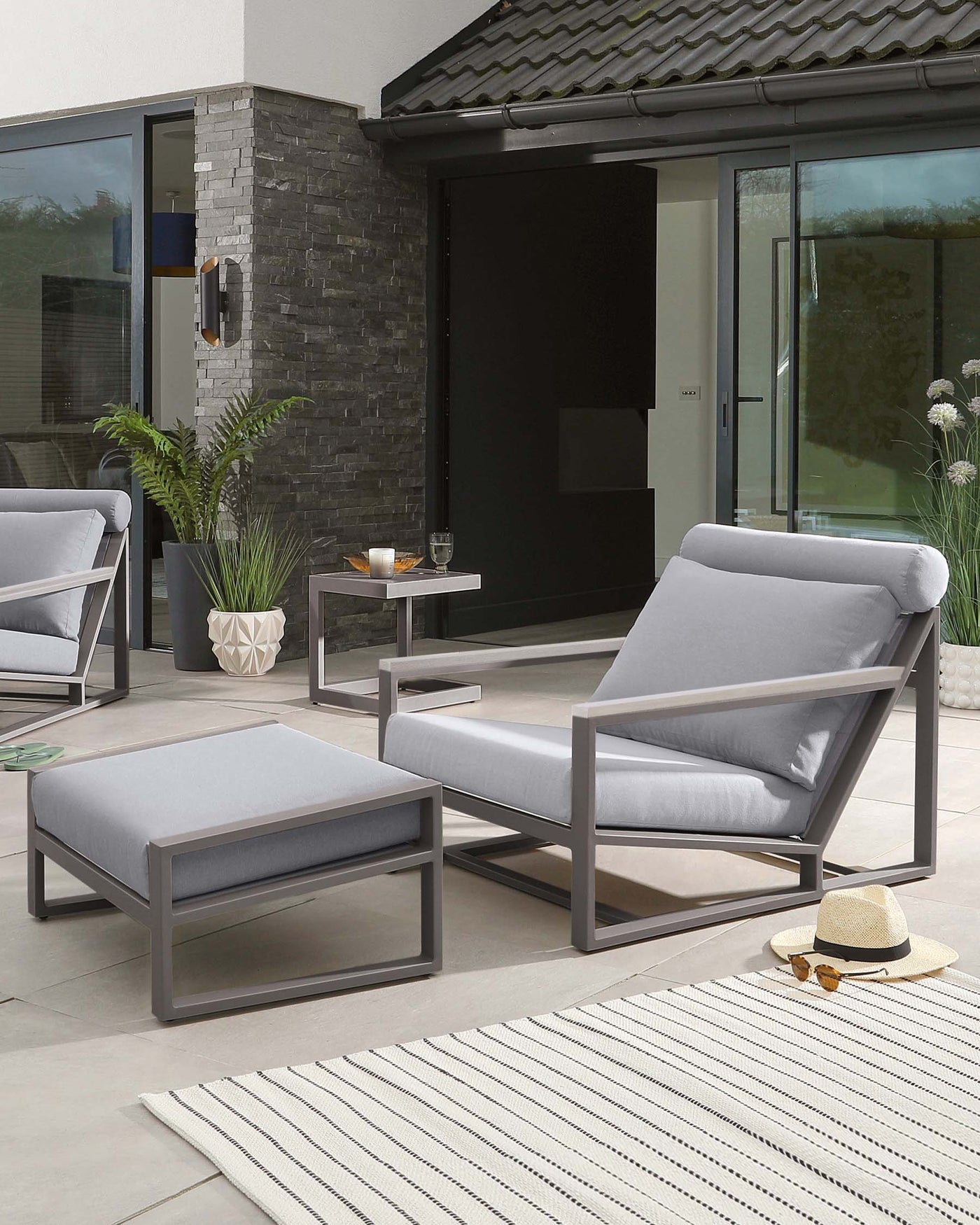 Modern outdoor furniture set with sleek grey metal frames, including two armchairs with comfortable light grey cushions, a matching rectangular side table, and a square ottoman also with a light grey cushion, arranged on a patio area with a striped area rug.
