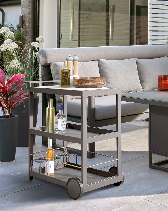 A modern outdoor rectangular metal side table with three shelves and wheels, in a matte grey finish. The top shelf holds decorative items and the lower shelves have bottles arranged neatly. In the background, a matching grey outdoor sofa with light grey cushions can be partially seen.