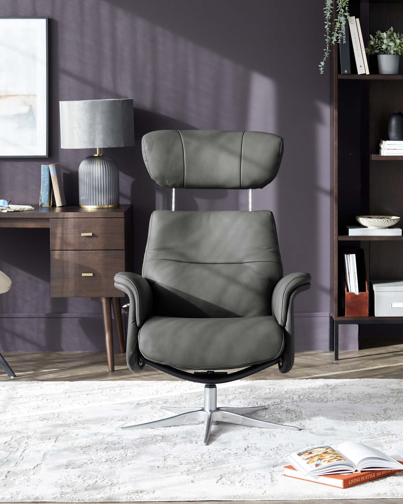 Modern black leather office chair with a high back, padded headrest, and armrests, featuring a swivel base with five castors. The chair is set in a stylish room with a dark brown wooden side table, a contemporary grey table lamp, bookshelves, and décor, complemented by a grey wall and plush white area rug.