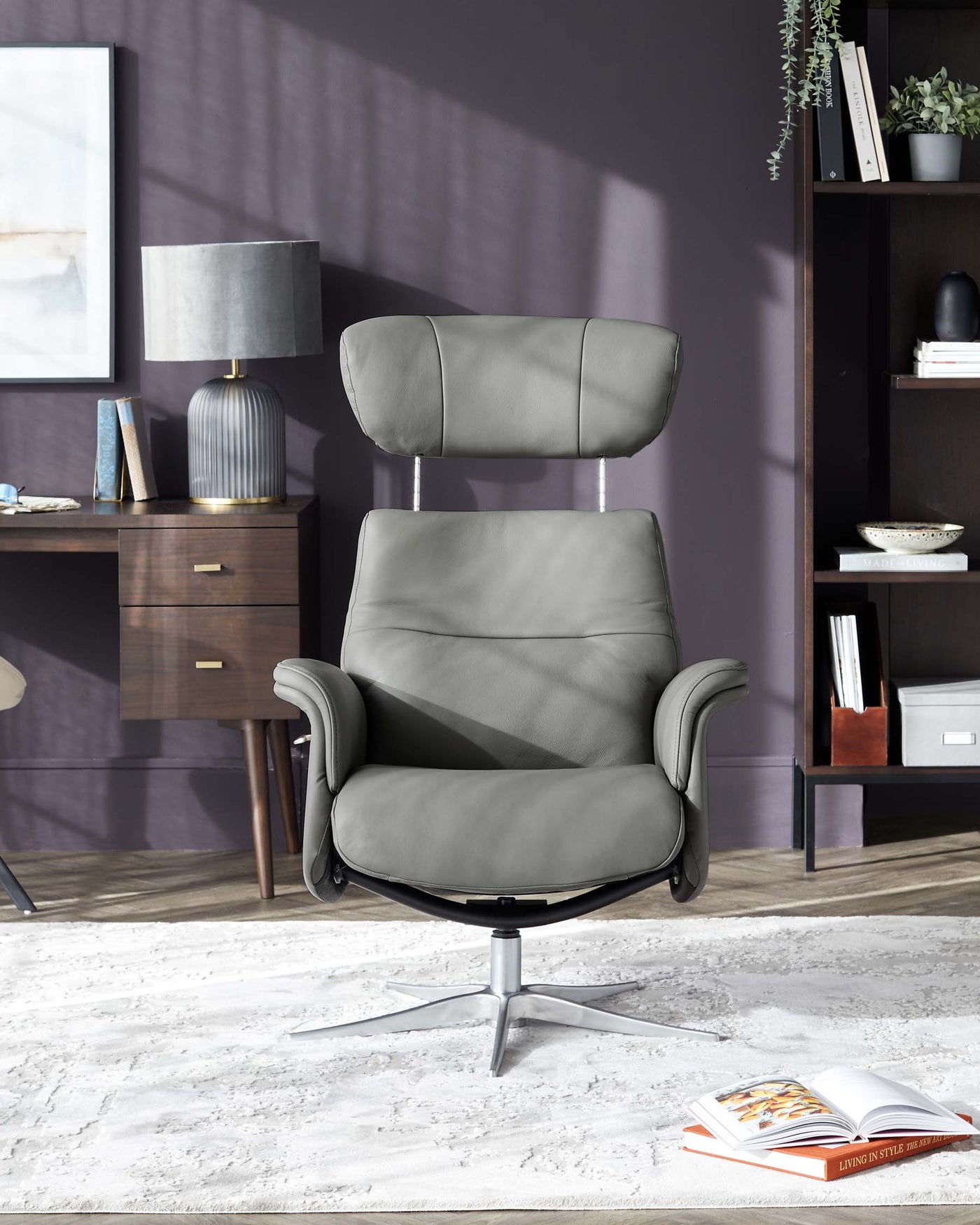 Modern charcoal-grey leather recliner with a high backrest and padded armrests featuring a swivel metal base, paired with a dark wood sideboard and shelving unit displaying decorative items and books, with a contemporary grey fabric table lamp on top of the sideboard.