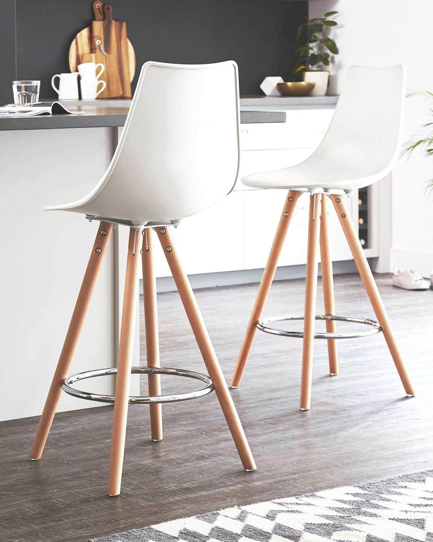 Two modern Scandinavian style bar stools with white plastic seats and wooden legs with metallic accents. Each stool features a low back design and a metal footrest circle. The stools are positioned in a bright kitchen with a subtle contemporary aesthetic.