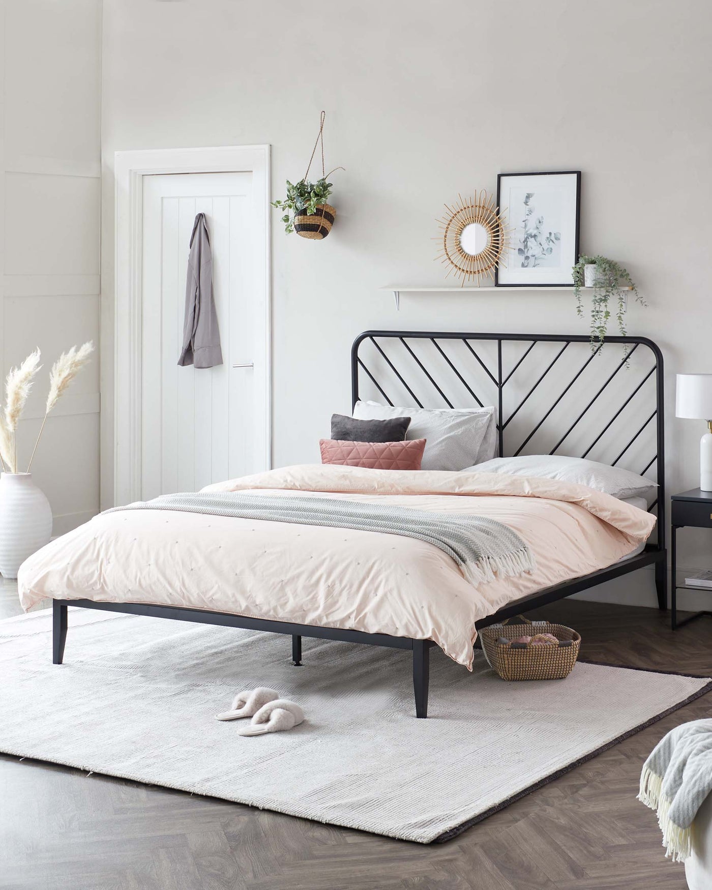 Contemporary minimalist style metal platform bed frame with a black finish featuring a headboard with a geometric pattern of intersecting lines. The bed is accessorized with a blush and grey bedding set consisting of a duvet cover, sheets, and assorted throw pillows. A light grey area rug is placed under the bed.
