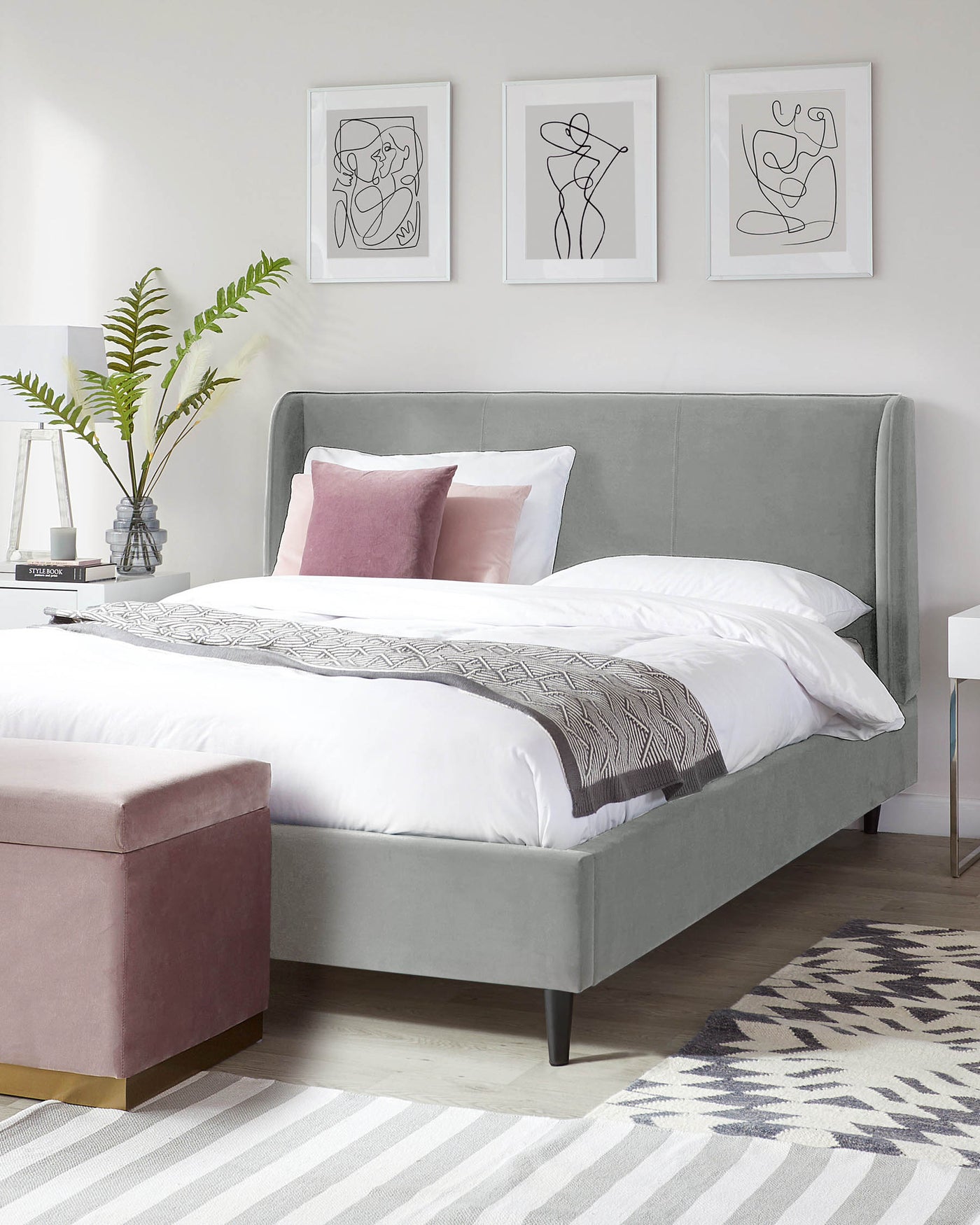 Elegant contemporary bedroom featuring a plush king-size bed with a curved, light grey upholstered headboard and frame on black tapered legs. At the foot of the bed sits a rectangular ottoman in a complementary dusky pink velvet with a metallic gold accent at the base. A minimalistic white side table is partially visible to the left. The space is accented by a grey and white striped area rug underneath.