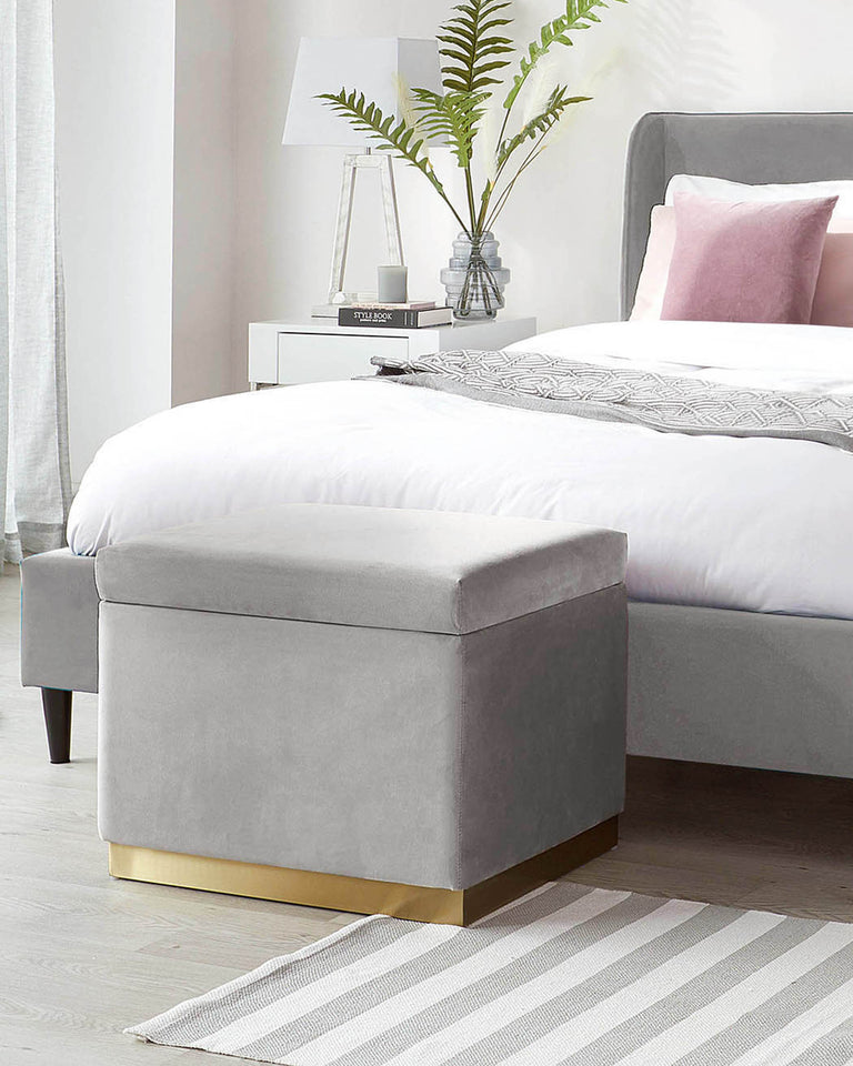 Contemporary bedroom furniture featuring a grey upholstered bed with a tall, slightly winged headboard, a white bedside table with a single drawer, and a modern clear glass lamp with a white shade. In the foreground, a grey fabric storage ottoman with a plush top and a shining gold metal base. The scene is complemented by light-coloured wooden flooring, a soft striped area rug, and fresh greenery for a touch of nature.
