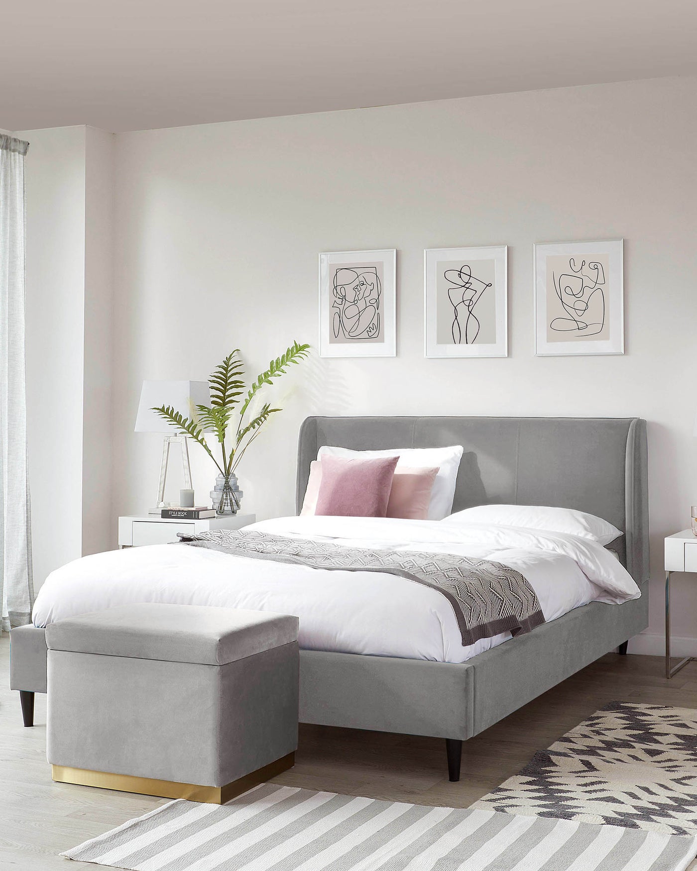 Upholstered platform bed with a high headboard in a soft grey fabric, accompanied by a matching grey storage ottoman with a gold-tone metal base.